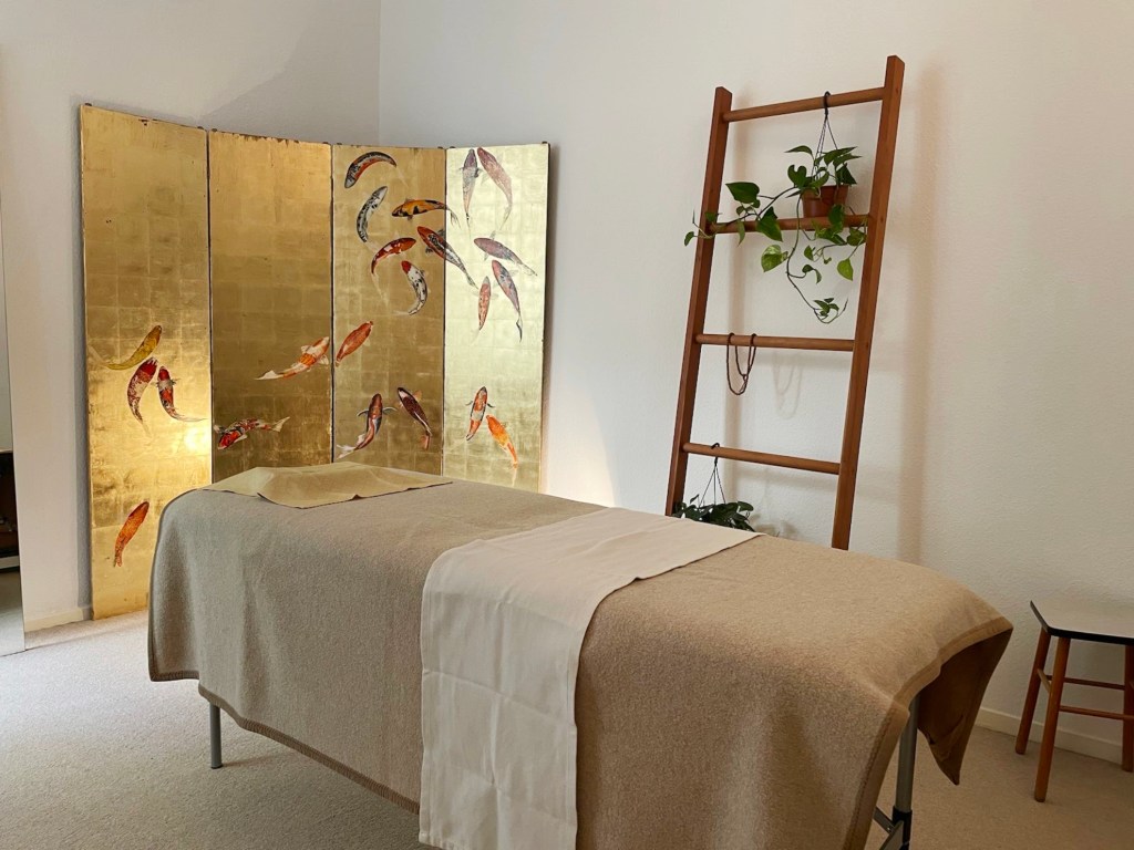 Hübsch arrangierter, gemütlicher Raum mit hellem Teppichboden, einer Massageliege, Pflanzen und buddhistischer Gebetskette auf einer hohen Pflanzenleiter und einem goldenem Paravent in der Ecke auf dem Goldfische zu sehen sind.